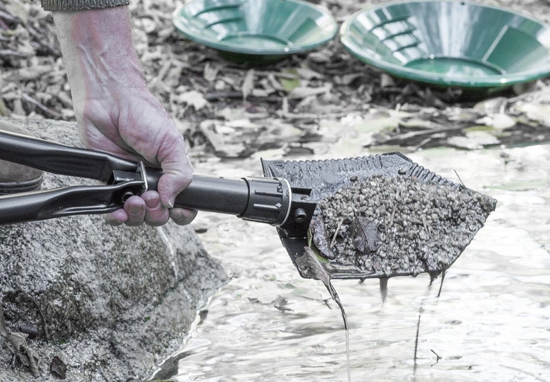May include: A black folding shovel with a metal handle is being used to scoop up gravel from a stream. The shovel is filled with gravel and water is dripping from the edge. Two green metal pans are in the background.