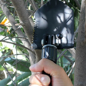 May include: A black folding shovel with a serrated edge and a black handle. The shovel is held in a hand.