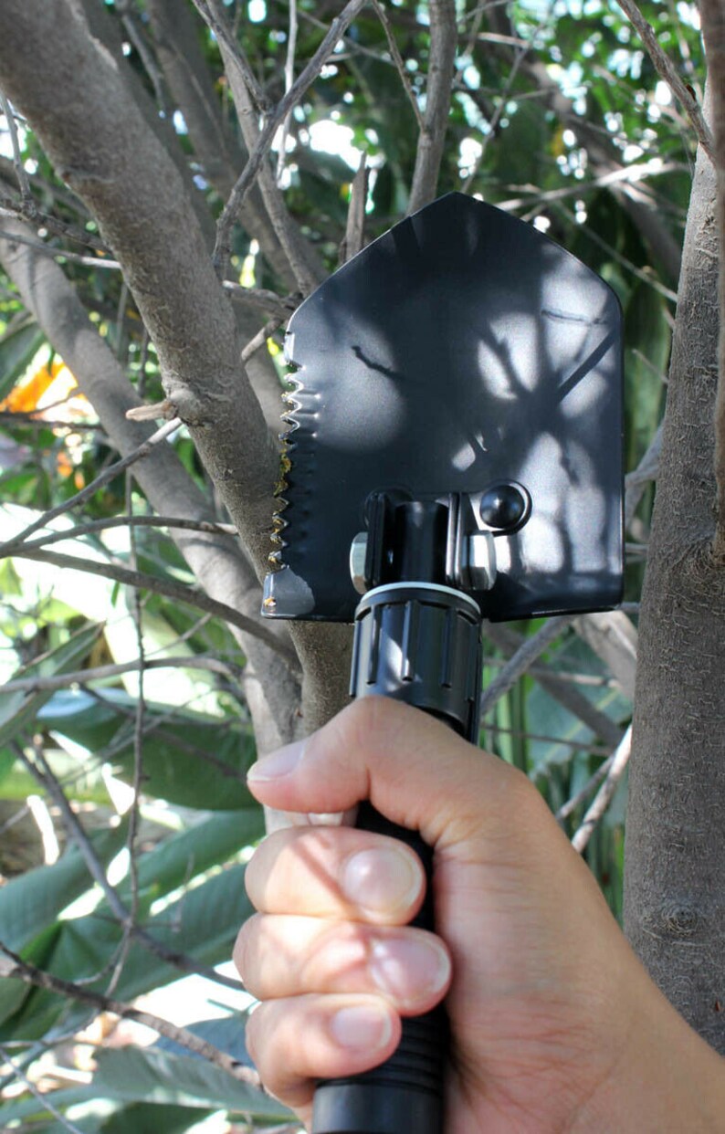 May include: A black folding shovel with a serrated edge and a black handle. The shovel is held in a hand.