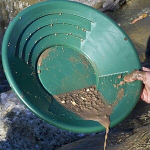 Puede incluir: Una batea de oro de pl&aacute;stico verde, llena de tierra y peque&ntilde;as piedras, se sostiene sobre el agua. La batea tiene crestas y una superficie texturizada. El contenido se est&aacute; derramando. Se ve una mano sosteniendo la batea.