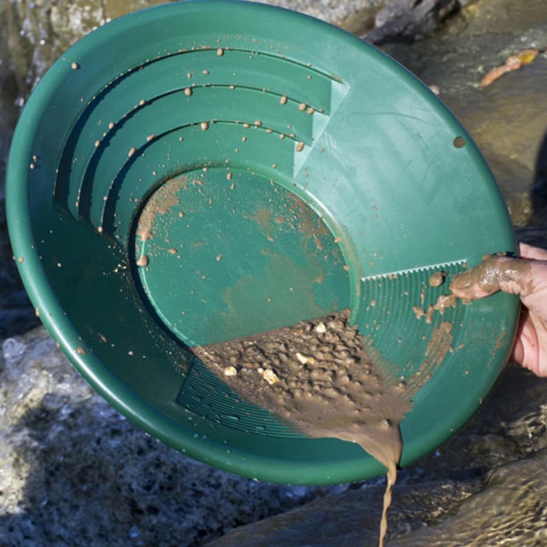 Puede incluir: Una batea de oro de pl&aacute;stico verde, llena de tierra y peque&ntilde;as piedras, se sostiene sobre el agua. La batea tiene crestas y una superficie texturizada. El contenido se est&aacute; derramando. Se ve una mano sosteniendo la batea.