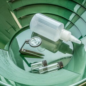 Puede incluir: Un conjunto de herramientas para la b&uacute;squeda de oro, que incluye una batea verde, una botella de pl&aacute;stico transparente, una lupa, unas pinzas y dos peque&ntilde;os viales de vidrio con tapas negras. Las herramientas est&aacute;n dispuestas sobre una superficie verde reflectante.