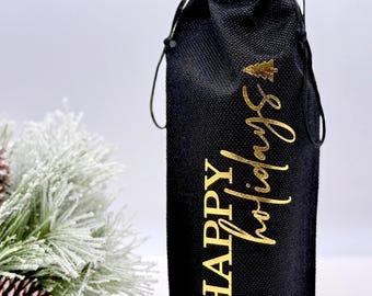 May include: Black wine bottle gift bag with gold lettering that reads "Happy Holidays" and a small Christmas tree icon. The bag is made of a textured material and has a drawstring closure. A sprig of frosted pine and pinecones are in the background.