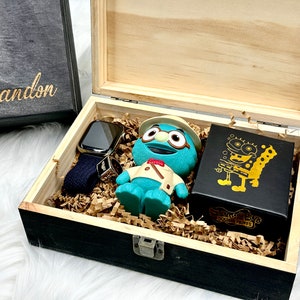May include: A wooden box with a black interior, filled with brown packing paper. The box contains a blue and green character toy, a black box with a gold character design, and a black and silver smartwatch with a blue band.