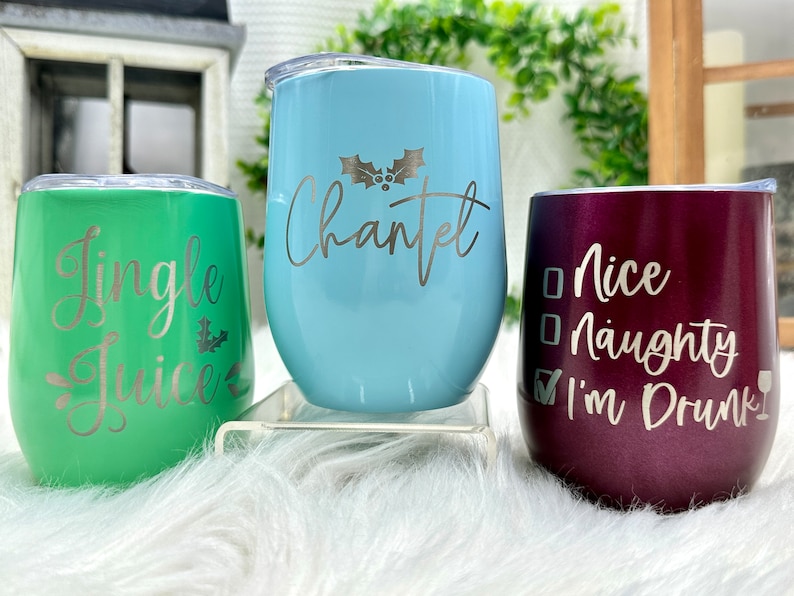 May include: Three stainless steel wine tumblers with Christmas-themed designs. The green tumbler says "Jingle Juice", the blue tumbler says "Chantel" with a holly sprig, and the purple tumbler says "Nice Naughty I'm Drunk" with a checkmark next to "I'm Drunk".