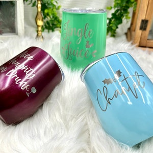 May include: Three stainless steel wine tumblers with different Christmas-themed designs. The tumblers are in burgundy, green, and light blue. The burgundy tumbler says "Nice Naughty or Drunk". The green tumbler says "Jingle Juice". The light blue tumbler says "Charlotte" with a holly sprig.
