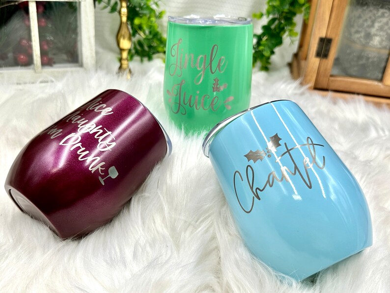 May include: Three stainless steel wine tumblers with different Christmas-themed designs. The tumblers are in burgundy, green, and light blue. The burgundy tumbler says "Nice Naughty or Drunk". The green tumbler says "Jingle Juice". The light blue tumbler says "Charlotte" with a holly sprig.
