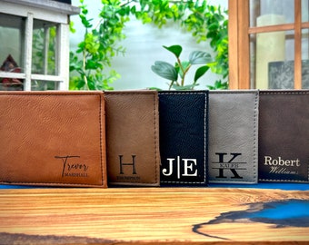 May include: Five personalized leather wallets in different colors. The wallets have different names and initials engraved on them. The wallets are arranged in a row on a wooden surface.