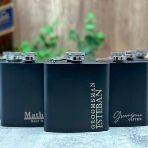 May include: A row of five black metal hip flasks with silver caps and personalized engravings. The flasks are engraved with the words "Groom's Man", "Esteban", "Math", "Best Man", and "Groomsman Oliver".