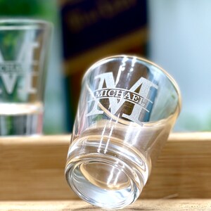 Op de afbeelding: Twee glazen shotglazen met een gegraveerd ontwerp. Het shotglas op de voorgrond heeft de letter "M" gegraveerd met de naam "MICHAEL" eronder.