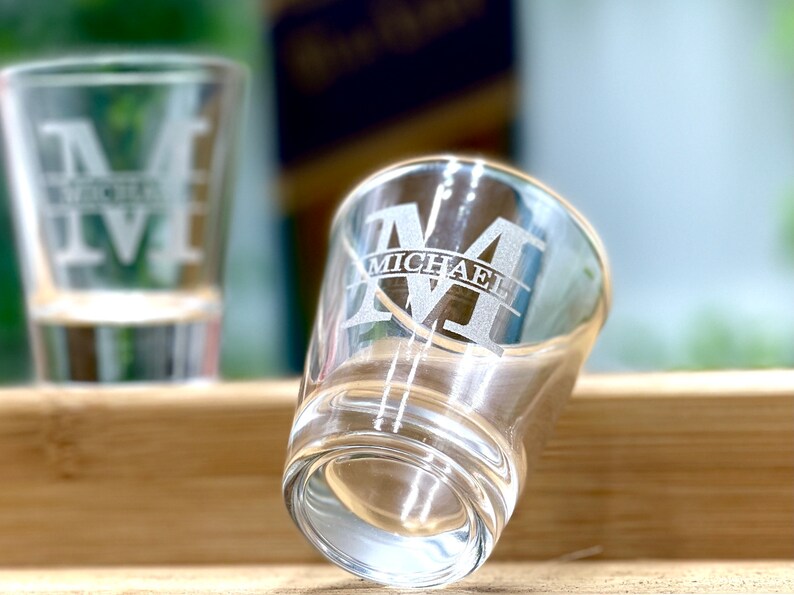 Op de afbeelding: Twee glazen shotglazen met een gegraveerd ontwerp. Het shotglas op de voorgrond heeft de letter "M" gegraveerd met de naam "MICHAEL" eronder.