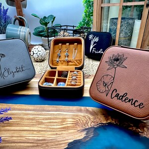Puede incluir: Tres cajas de joyer&iacute;a personalizadas con diferentes dise&ntilde;os florales y nombres. Las cajas son grises, marrones y negras. La caja gris tiene un dise&ntilde;o de margarita y el nombre "Elizabeth" grabado en ella. La caja marr&oacute;n tiene un dise&ntilde;o de flor de loto y el nombre "Cadence" grabado en ella. La caja negra tiene un peque&ntilde;o dise&ntilde;o floral y el nombre "Frida" grabado en ella.