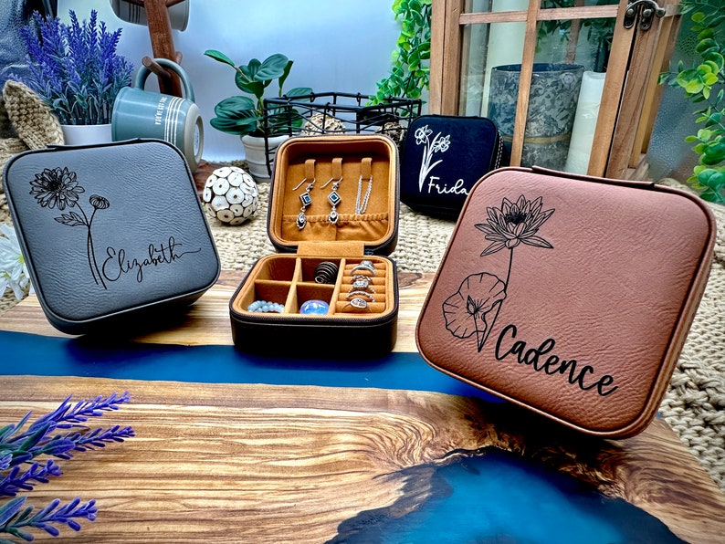 Puede incluir: Tres cajas de joyer&iacute;a personalizadas con diferentes dise&ntilde;os florales y nombres. Las cajas son grises, marrones y negras. La caja gris tiene un dise&ntilde;o de margarita y el nombre "Elizabeth" grabado en ella. La caja marr&oacute;n tiene un dise&ntilde;o de flor de loto y el nombre "Cadence" grabado en ella. La caja negra tiene un peque&ntilde;o dise&ntilde;o floral y el nombre "Frida" grabado en ella.