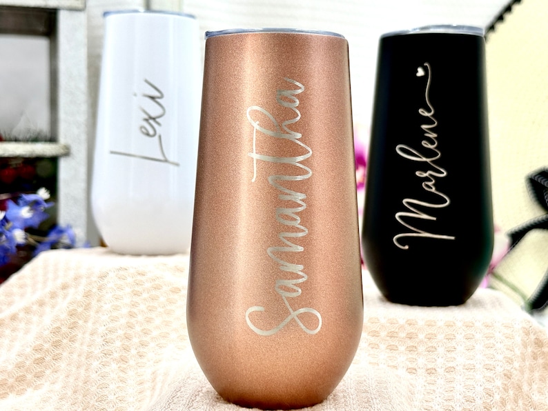May include: Three stemless tumblers with lids, featuring a metallic rose gold finish with the name Samantha, a matte white finish with the name Lexi, and a matte black finish with the name Marlene, all engraved in a cursive script font.