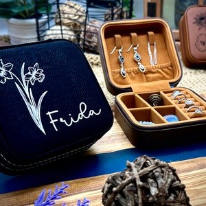 Puede incluir: Dos cajas de joyer&iacute;a negras y marrones con un dise&ntilde;o floral y el nombre "Frida" en una de las cajas. La otra caja est&aacute; abierta y tiene un interior marr&oacute;n con compartimentos para anillos y pendientes.