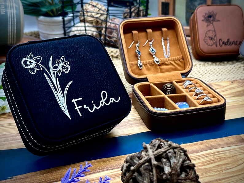 Puede incluir: Dos cajas de joyer&iacute;a negras y marrones con un dise&ntilde;o floral y el nombre "Frida" en una de las cajas. La otra caja est&aacute; abierta y tiene un interior marr&oacute;n con compartimentos para anillos y pendientes.