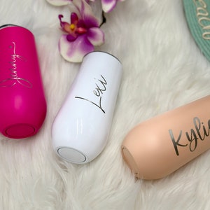 May include: Three stainless steel stemless champagne flutes in hot pink, white, and beige. Each tumbler features a custom name in elegant script lettering: Jenny on the pink, Lexi on the white, and Kylie on the beige. Each cup has a black lid.