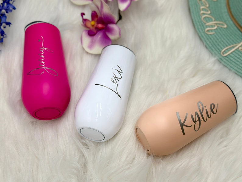 May include: Three stainless steel stemless champagne flutes in hot pink, white, and beige. Each tumbler features a custom name in elegant script lettering: Jenny on the pink, Lexi on the white, and Kylie on the beige. Each cup has a black lid.
