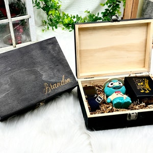 May include: A black wooden box with the name "Brandon" engraved on the lid. The box is open and contains a watch, a blue toy, and a black box with gold lettering.