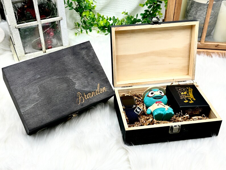 May include: A black wooden box with the name "Brandon" engraved on the lid. The box is open and contains a watch, a blue toy, and a black box with gold lettering.