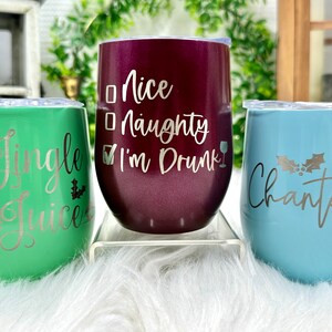 May include: Three stainless steel wine tumblers with festive Christmas designs. The green tumbler says "Jingle Juice", the burgundy tumbler says "Nice Naughty I'm Drunk", and the blue tumbler says "Chantel" with a holly sprig.