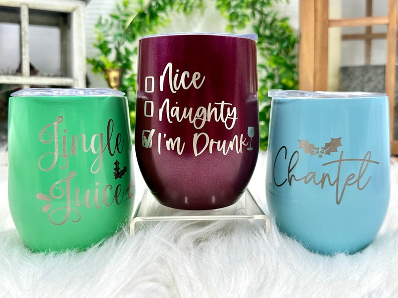 May include: Three stainless steel wine tumblers with festive Christmas designs. The green tumbler says "Jingle Juice", the burgundy tumbler says "Nice Naughty I'm Drunk", and the blue tumbler says "Chantel" with a holly sprig.