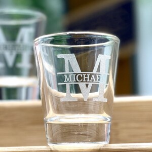 Op de afbeelding: Twee heldere glazen shotglazen met een gepersonaliseerd ontwerp. Het shotglas rechts heeft de letter "M" en de naam "MICHAEL" gegraveerd in het glas.