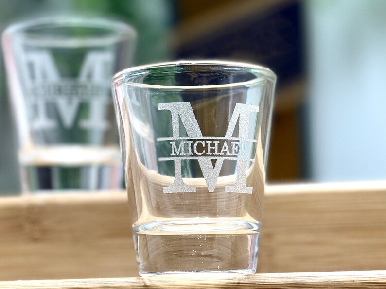 Op de afbeelding: Twee heldere glazen shotglazen met een gepersonaliseerd ontwerp. Het shotglas rechts heeft de letter "M" en de naam "MICHAEL" gegraveerd in het glas.
