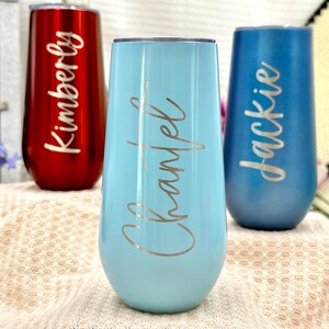 May include: Three stainless steel stemless champagne flutes with lids. The left tumbler is metallic red with the name Kimberly, the center is light blue with the name Chantel, and the right is matte blue with the name Jackie engraved in script.