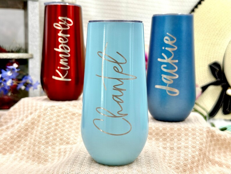 May include: Three stainless steel stemless champagne flutes with lids. The left tumbler is metallic red with the name Kimberly, the center is light blue with the name Chantel, and the right is matte blue with the name Jackie engraved in script.