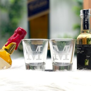 Op de afbeelding: Twee heldere glazen shotglazen met de letter "M" gegraveerd op de voorkant. De glazen staan op een wit oppervlak naast een klein flesje tequila. De fles heeft een zwart etiket met de tekst "Tequila 100% de Agave" en "Al", en een rode waszegel.