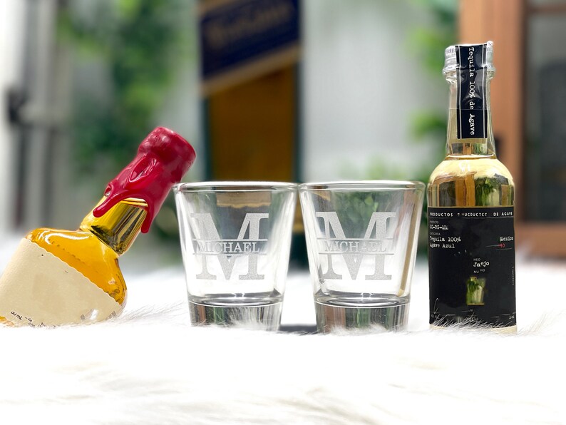 Op de afbeelding: Twee heldere glazen shotglazen met de letter "M" gegraveerd op de voorkant. De glazen staan op een wit oppervlak naast een klein flesje tequila. De fles heeft een zwart etiket met de tekst "Tequila 100% de Agave" en "Al", en een rode waszegel.