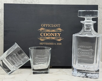 May include: A clear glass decanter and two matching square whiskey glasses, each etched with the text "OFFICIANT COONEY" and "SEPTEMBER 8, 2026". Behind them sits a black wooden box featuring the same text and date in gold lettering.