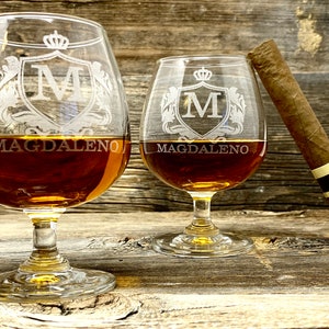May include: Two brandy snifters filled with amber liquid, engraved with a crown and the letter "M" inside a crest, and the name "Magdaleno". A cigar rests against one glass on a wooden surface.