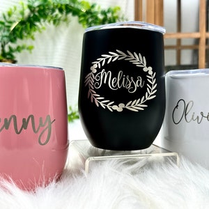 May include: Three stainless steel tumblers with personalized Christmas-themed designs. The pink tumbler has a Santa hat and the name "Jenny". The black tumbler has a wreath and the name "Melissa". The white tumbler has ornaments and the name "Olivia".