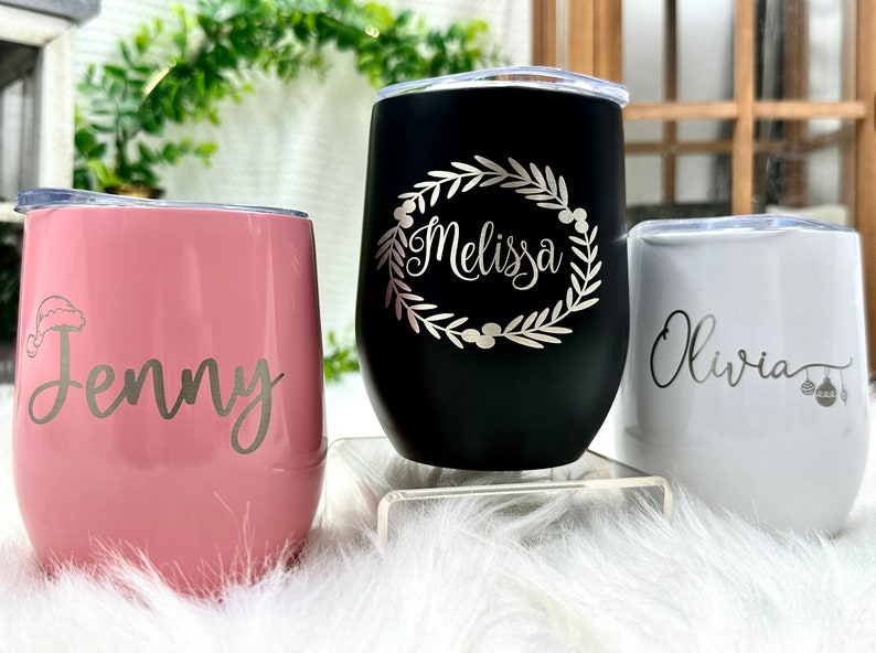 May include: Three stainless steel tumblers with personalized Christmas-themed designs. The pink tumbler has a Santa hat and the name "Jenny". The black tumbler has a wreath and the name "Melissa". The white tumbler has ornaments and the name "Olivia".