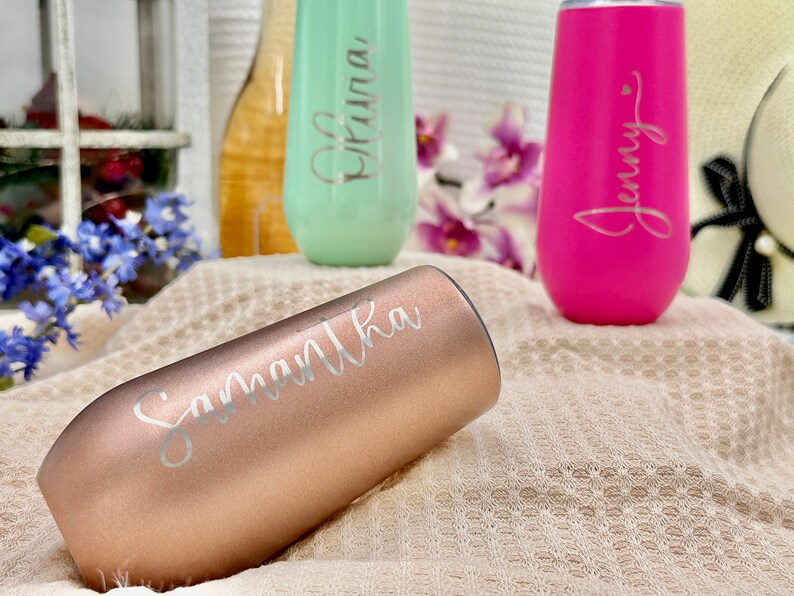 May include: Three stainless steel stemless champagne flutes with a matte finish. One is rose gold with the name Samantha, one is mint green with the name Olivia, and one is hot pink with the name Jenny. Each flute features custom script lettering.