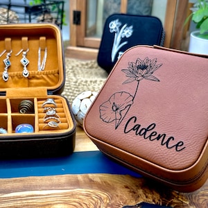 Puede incluir: Dos cajas de joyer&iacute;a de cuero marr&oacute;n con cremalleras. Una caja tiene un dise&ntilde;o floral y el nombre "Cadence" grabado en ella. La otra caja est&aacute; abierta y tiene compartimentos para anillos, pendientes y una pulsera.