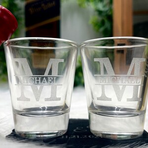 Op de afbeelding: Twee heldere glazen shotglazen met de naam "Michael" gegraveerd in een groot, vet lettertype. De glazen staan op een zwart oppervlak.