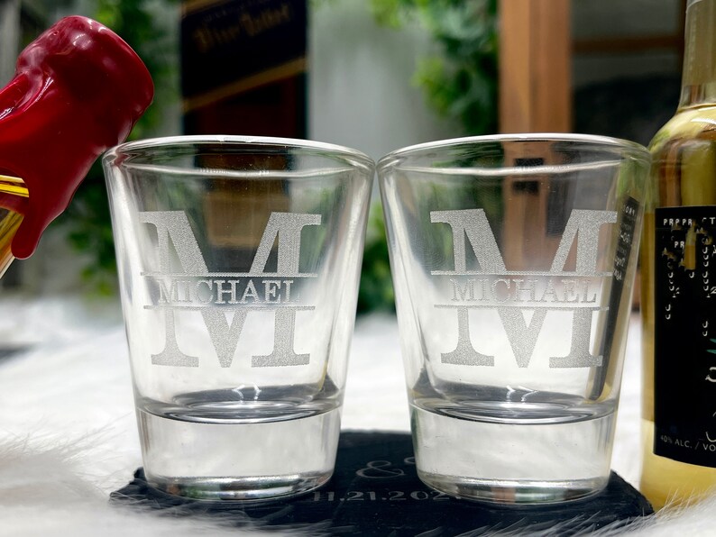 Op de afbeelding: Twee heldere glazen shotglazen met de naam "Michael" gegraveerd in een groot, vet lettertype. De glazen staan op een zwart oppervlak.