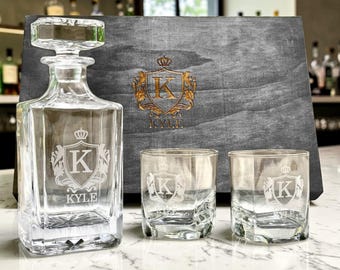 May include: A clear glass decanter with a matching stopper and two matching glasses, each etched with a crest featuring the letter K and the name KYLE. Behind them sits a dark gray wooden box with a gold-colored crest and the name KYLE.