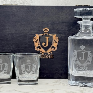 May include: A clear glass decanter with a matching stopper and two square glasses, each etched with a crest featuring the letter J and the name JESSE. The set is displayed in front of a black wooden box with a gold crest and the name JESSE.