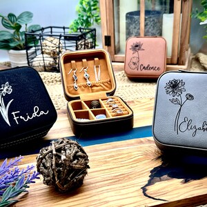 Puede incluir: Tres peque&ntilde;as cajas cuadradas de joyer&iacute;a con dise&ntilde;os florales y nombres personalizados. Las cajas son negras, grises y marrones. La caja negra tiene un dise&ntilde;o de narciso blanco y el nombre "Frida" en ella. La caja gris tiene un dise&ntilde;o de crisantemo blanco y el nombre "Elizabeth" en ella. La caja marr&oacute;n tiene un dise&ntilde;o de loto blanco y el nombre "Cadence" en ella.
