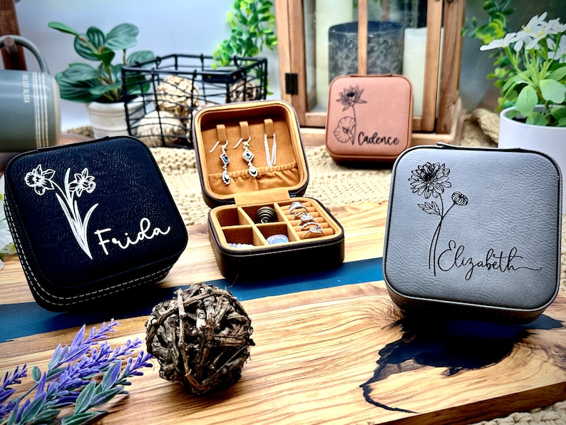 Puede incluir: Tres peque&ntilde;as cajas cuadradas de joyer&iacute;a con dise&ntilde;os florales y nombres personalizados. Las cajas son negras, grises y marrones. La caja negra tiene un dise&ntilde;o de narciso blanco y el nombre "Frida" en ella. La caja gris tiene un dise&ntilde;o de crisantemo blanco y el nombre "Elizabeth" en ella. La caja marr&oacute;n tiene un dise&ntilde;o de loto blanco y el nombre "Cadence" en ella.