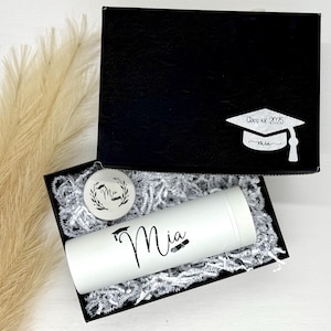 May include: A graduation gift set in a black box, including a white tumbler, ornament, and box lid with a graduation cap design. The tumbler and ornament are personalized with the name "Mia". The box is filled with white paper filler.