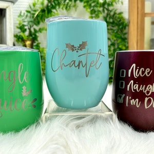 May include: Three stainless steel wine tumblers with Christmas-themed designs. The green tumbler says "Jingle Juice", the blue tumbler says "Chantel", and the burgundy tumbler says "Nice Naughty I'm Drunk".