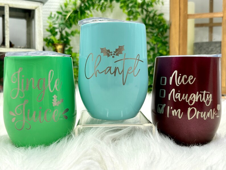 May include: Three stainless steel wine tumblers with Christmas-themed designs. The green tumbler says "Jingle Juice", the blue tumbler says "Chantel", and the burgundy tumbler says "Nice Naughty I'm Drunk".