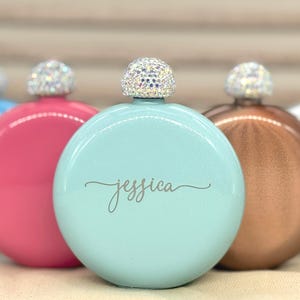May include: A collection of round, metallic-finish flasks in various colours including light blue, pink, mint green, copper, and white. Each flask features a screw-top cap encrusted with clear, sparkling rhinestones. The mint green flask has the name jessica engraved in a cursive script on the front.