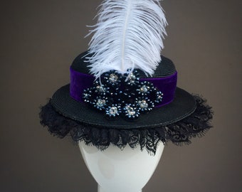 May include: A black hat with a wide brim, adorned with a purple velvet band and a cluster of sparkling embellishments. A large white feather and black lace trim add to the hat's elegant design.