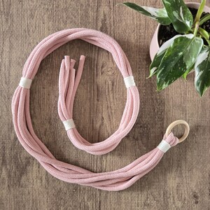 May include: A pink macrame plant hanger with a wooden ring. The hanger is made of thick cotton rope and has a white trim.